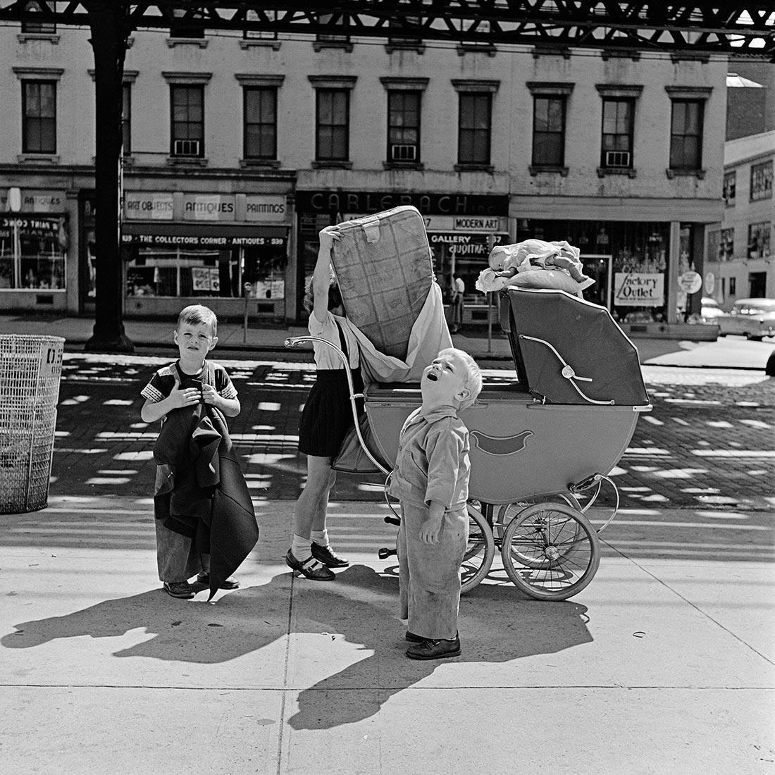 Streetscene New York 1950 ' s
Building in the background. Two children looking up and sideways from t the camera . A female loading a large box upon a child-pram